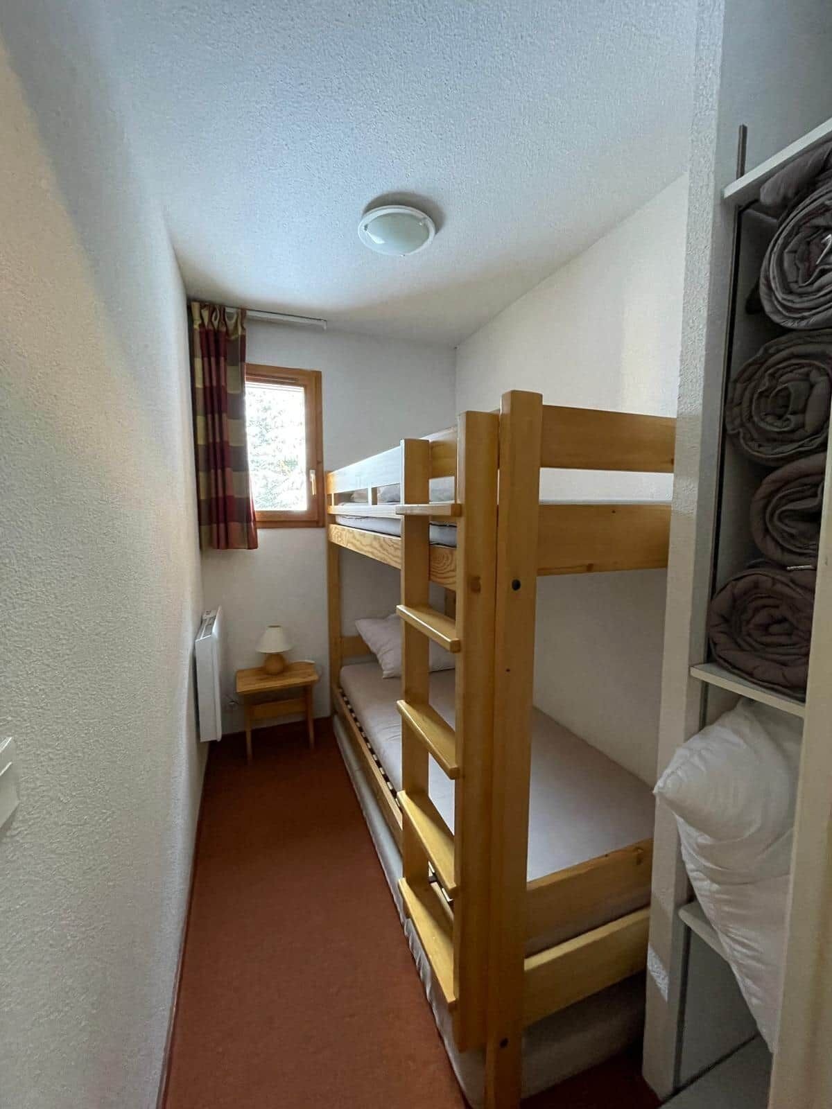A compact room with bunk beds, a small table, a window with curtains, and a radiator.