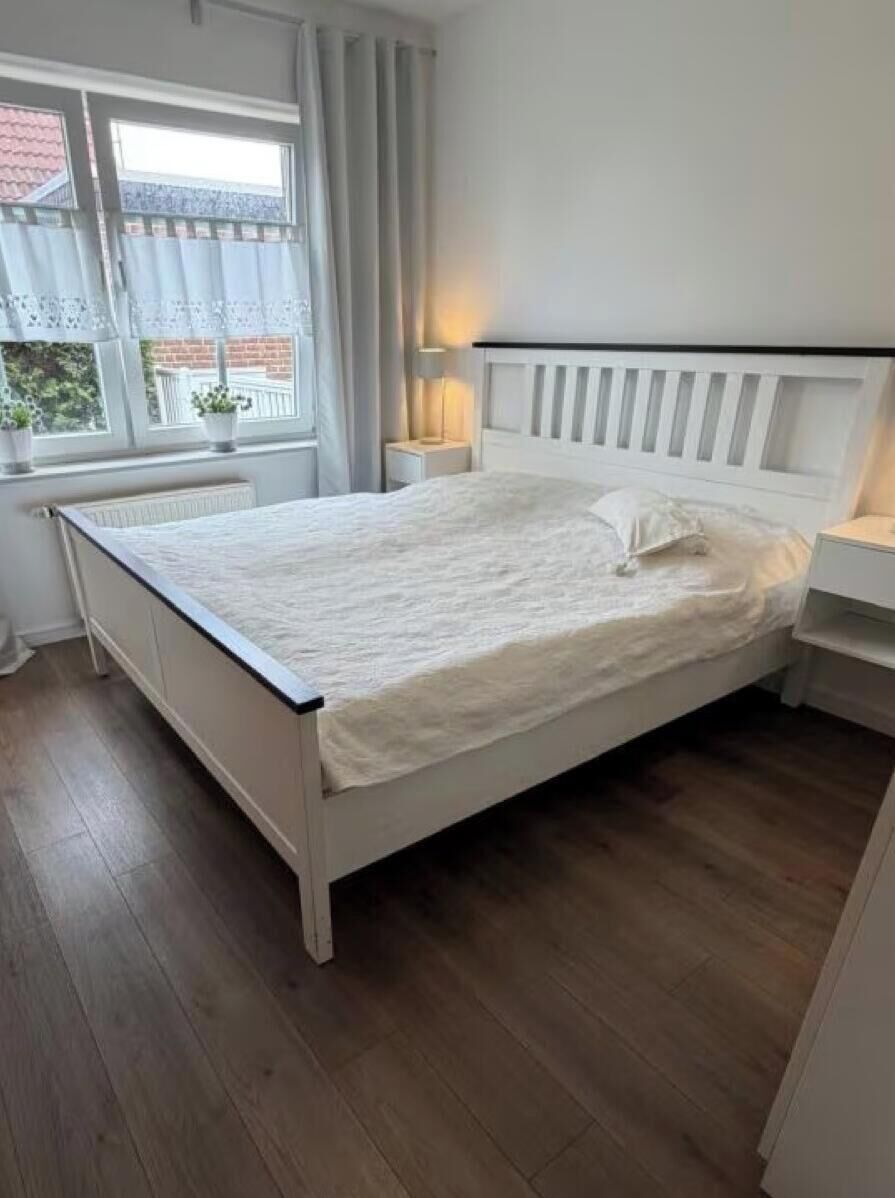 Une chambre à coucher avec un lit blanc, une table de chevet, une lampe et une fenêtre avec des rideaux.