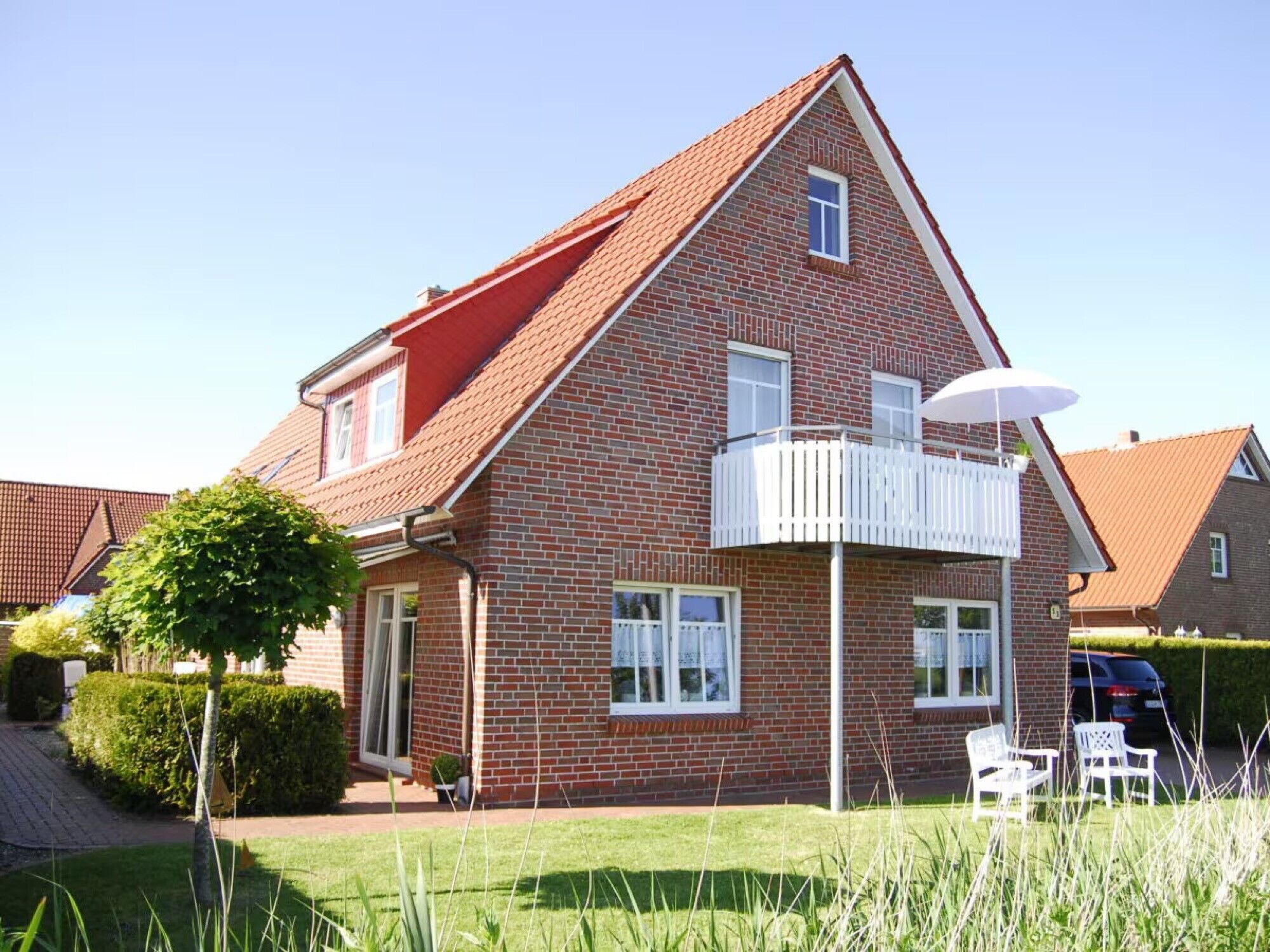 Ein zweistöckiges Backsteinhäuschen mit rotem Dach, einem Balkon mit weißer Brüstung und einem kleinen Garten mit Baum und Rasen.