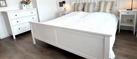 A bedroom with a white bed, a nightstand with a lamp, a dresser, and a wall with framed pictures.