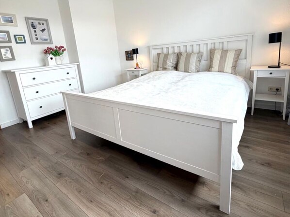 A bedroom with a white bed, a nightstand with a lamp, a dresser, and a wall with framed pictures.