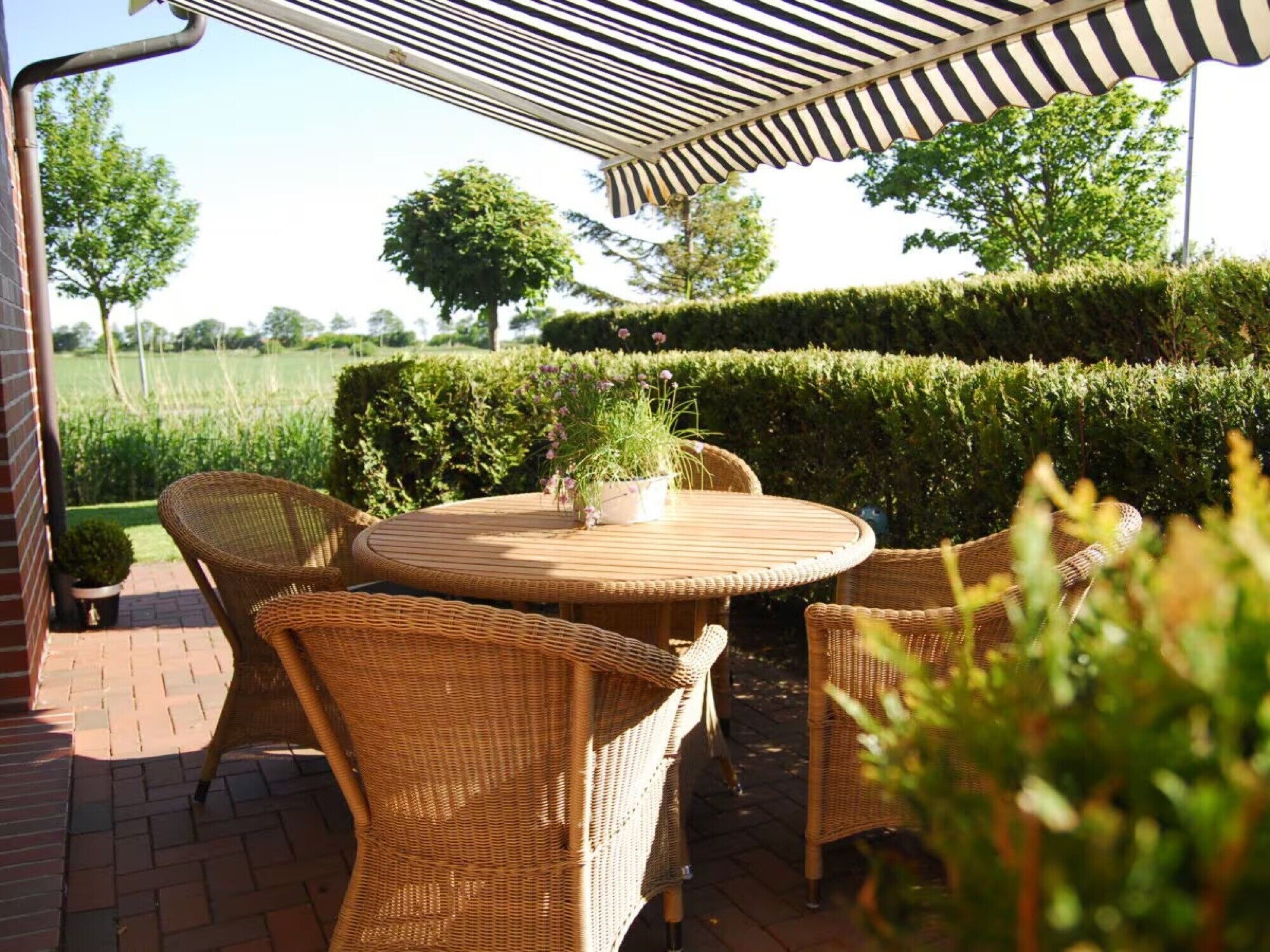 Eine Terrasse im Freien mit Korbmöbeln, einem runden Tisch und einem gestreiften Sonnensegel.