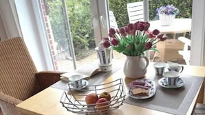 A dining area with a table set for tea, a wicker chair, and a view of a garden through large windows. - Apartment 'Anna Nordsee' with Private Terrace, Private Garden and Wi-Fi (Krummhörn)