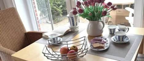 A dining area with a table set for tea, a wicker chair, and a view of a garden through large windows.