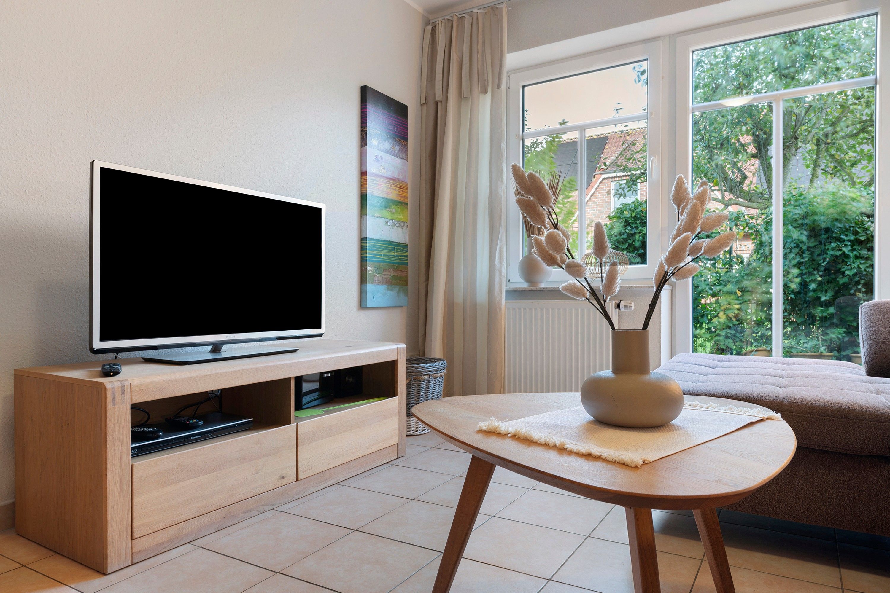 Un salon moderne avec un téléviseur à écran plat posé sur un meuble en bois, un vase contenant des plantes séchées sur une petite table, et une vue sur la verdure à travers de grandes fenêtres.