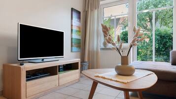 Un salon moderne avec un téléviseur à écran plat posé sur un meuble en bois, un vase contenant des plantes séchées sur une petite table, et une vue sur la verdure à travers de grandes fenêtres.