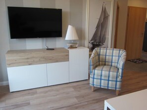 Ein modernes Wohnzimmer mit einem an der Wand montierten Flachbildfernseher, einer weißen Kommode mit Holzplatte, einer weißen Stehlampe, einem karierten Sessel und einer Holztür.