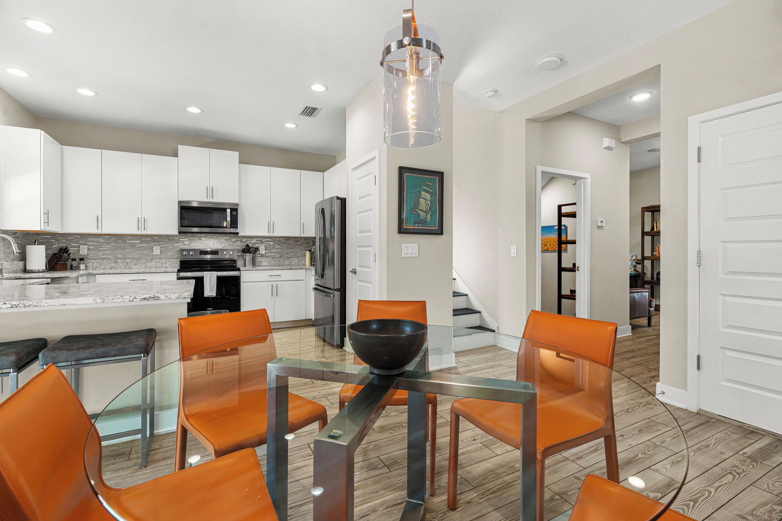 A modern kitchen with white cabinets, stainless steel appliances, and a glass dining table with orange chairs.