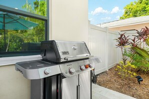 A Weber Genesis gas grill with four burners, positioned on a patio with a view of a house and a garden. - Mins to Beaches/ Downtown, Heated Pool, Arlington Park (Sarasota)