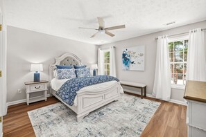 Un dormitorio con cama, mesitas de noche, un banco, ventilador de techo y ventana con cortinas.