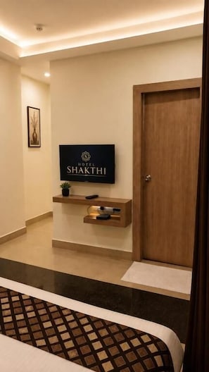Zona de recepción de un hotel con un televisor de pantalla plana montado en la pared, una puerta de madera y un estante pequeño con una planta y un control remoto.