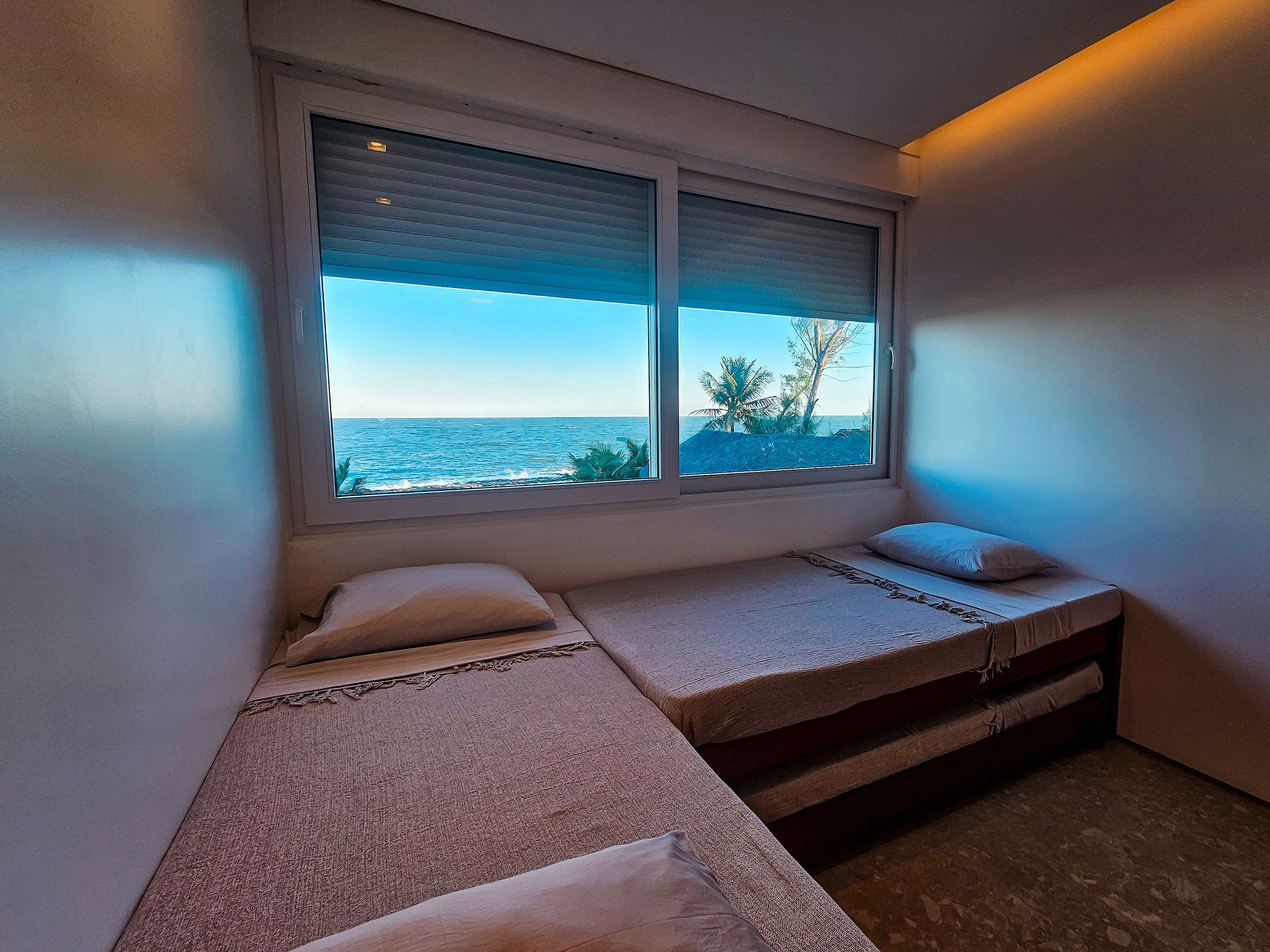 A room with two beds, a window with a view of the sea, and a small table.