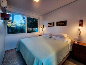 A bedroom with a bed, a nightstand, a wall-mounted lamp, and a window with a view of greenery. - Casa Geller (Ipojuca)
