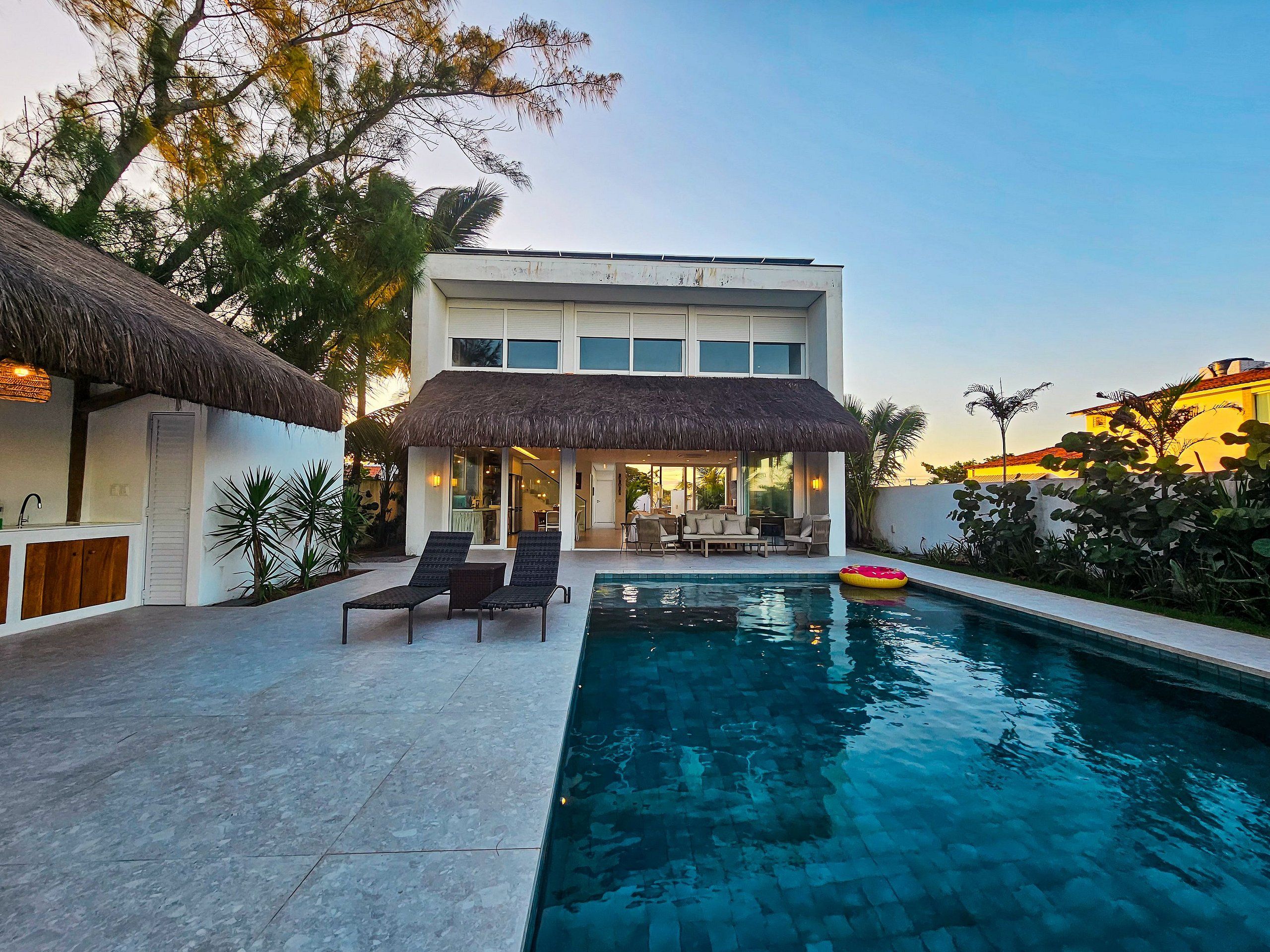 A modern building with a pool, lounge chairs, and a thatched-roof structure.