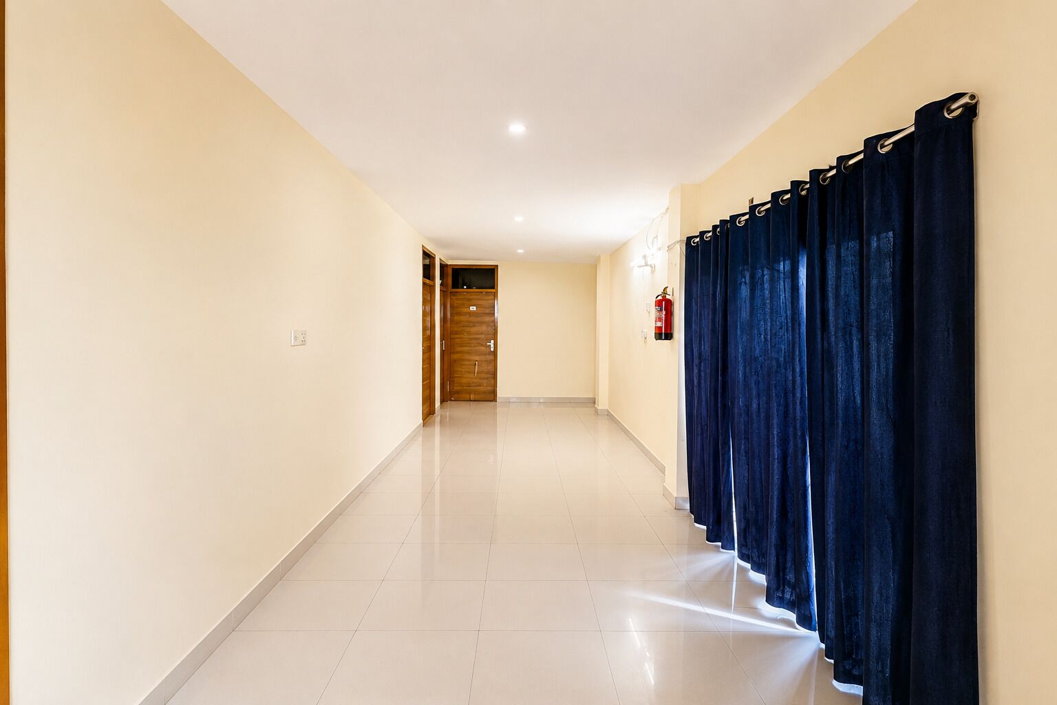 Um corredor com um extintor preso na parede, uma porta de madeira ao fundo e uma fileira de cortinas escuras à direita.