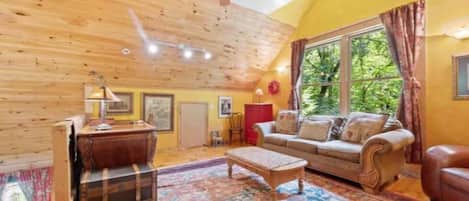 A cozy living room with a wooden ceiling, a sofa set, a coffee table, and a large window with curtains.