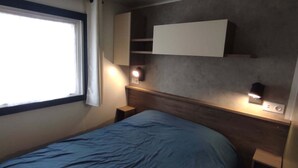 A room with a bed, a window, a wall-mounted shelf, and bedside lamps. - Siblu 4* Capbreton 3-Bedroom Solar-Powered Mobile Home — Available (Capbreton)