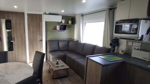 A compact living space with a sofa, a small table, a kitchen area with a microwave, and a window. - Siblu 4* Capbreton 3-Bedroom Solar-Powered Mobile Home — Available (Capbreton)