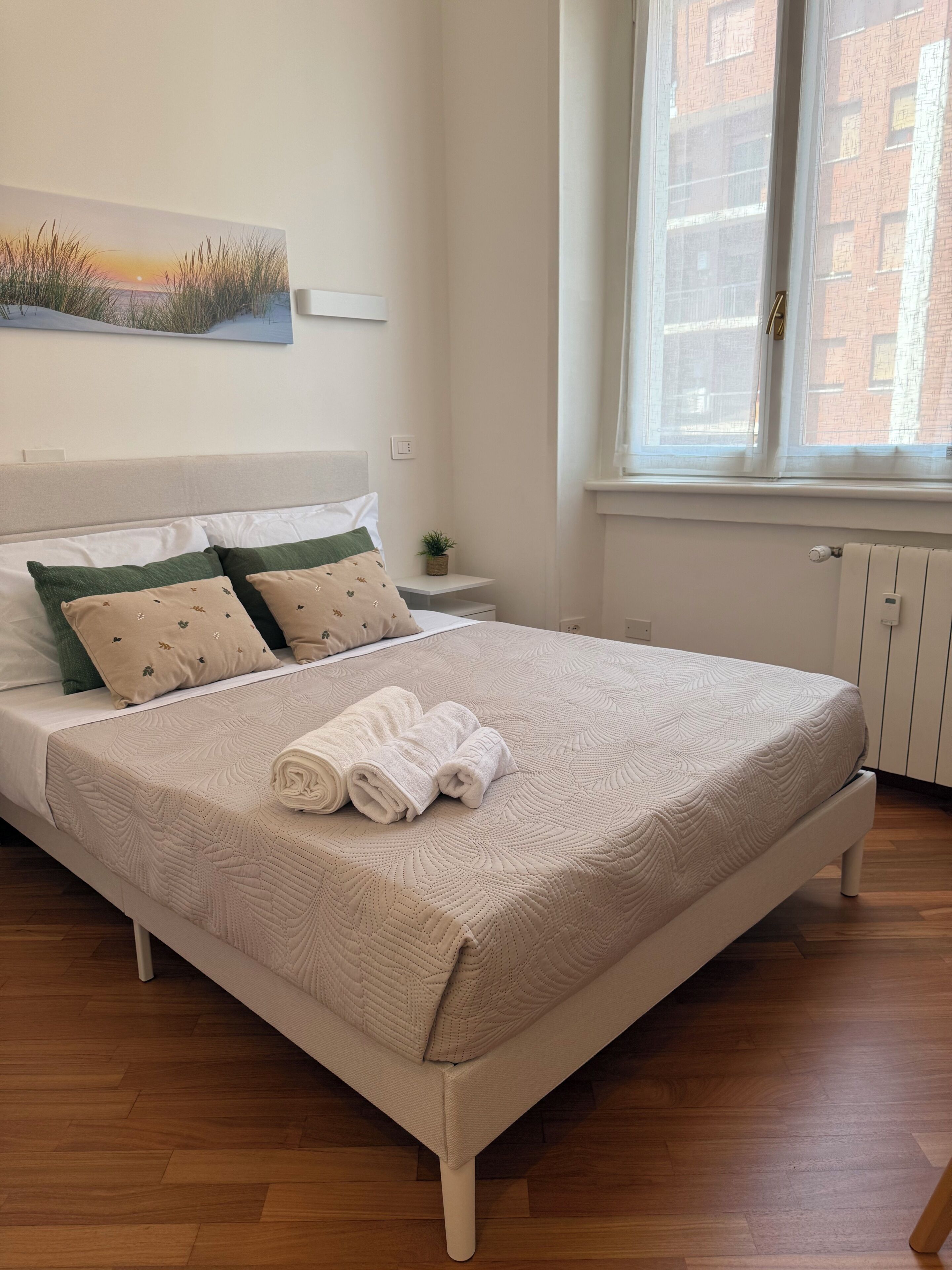Una cama bien hecha con colcha, almohadas y toallas enrolladas. Hay una mesita de noche con una planta, una ventana con vistas a edificios, y un cuadro colgado en la pared.