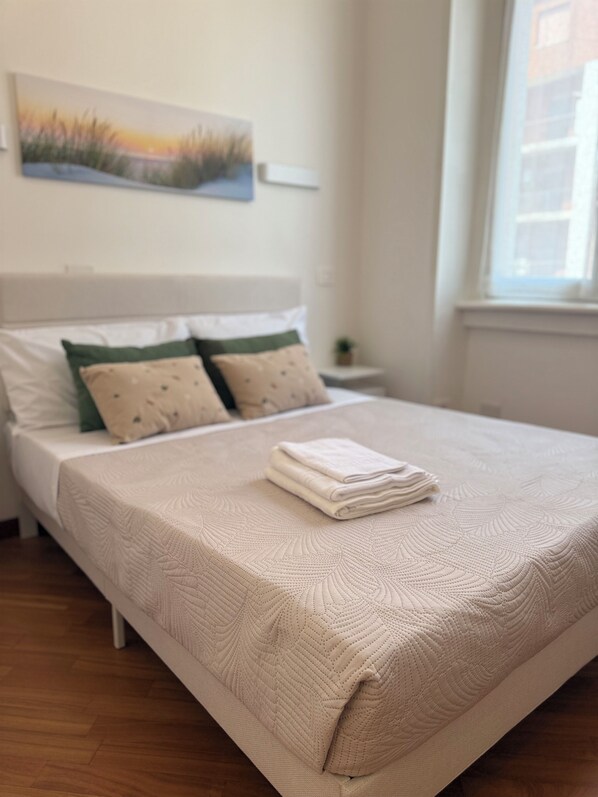Una cama bien hecha con cubrecama y almohadas con textura, una mesita de noche con lámpara y un cuadro en la pared.