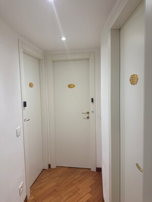 Un pasillo con dos puertas blancas, cada una con una placa dorada. Hay un aplique en el techo y un interruptor de luz en la pared.