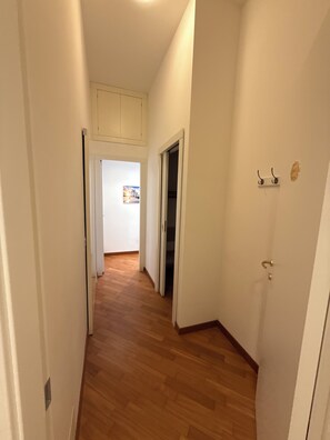 Um corredor com piso de madeira, paredes brancas e uma porta que dá para um quarto com uma pintura da paisagem urbana.