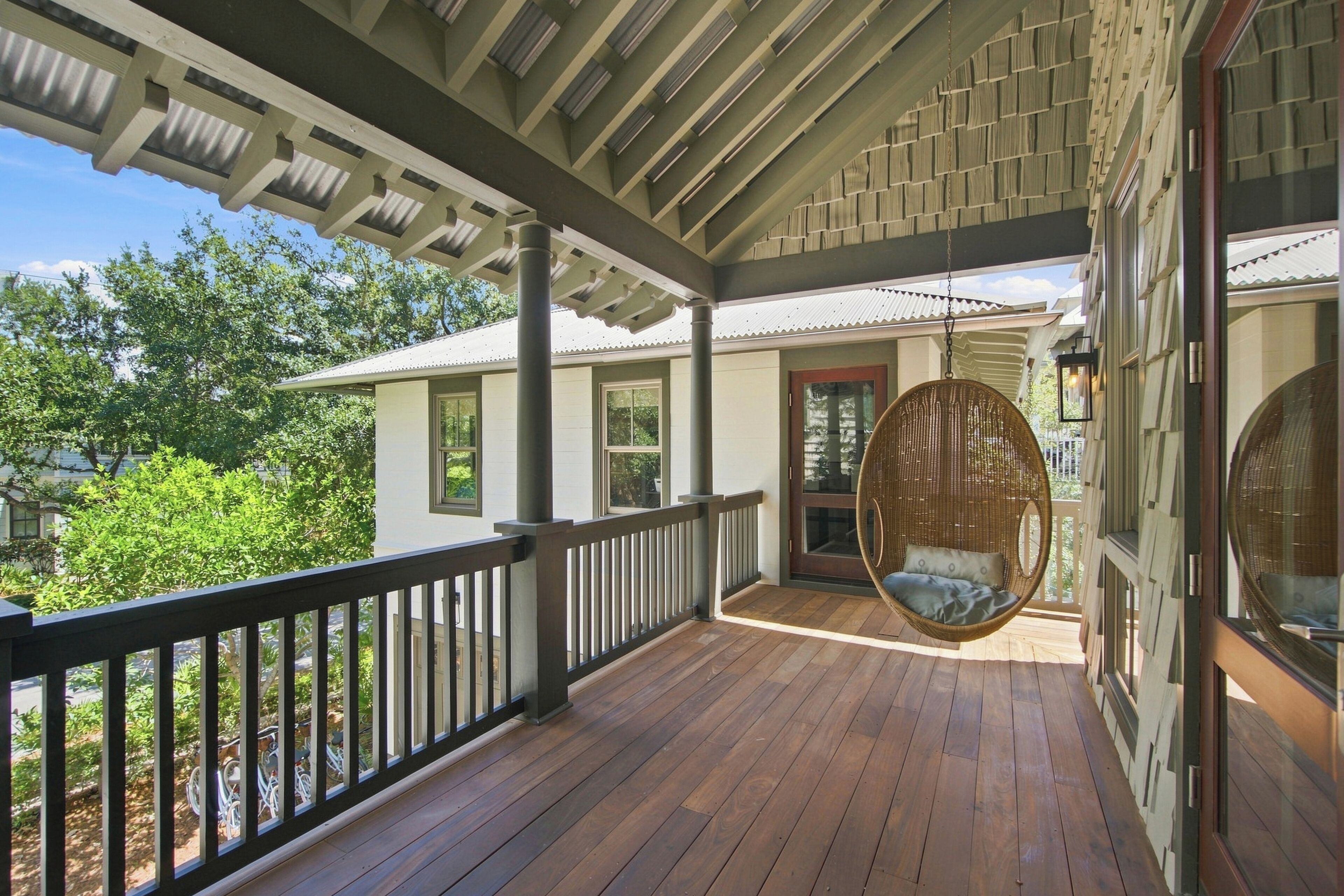 Une véranda couverte avec un fauteuil suspendu, une balustrade en bois et une vue sur une maison avec une porte et des fenêtres.