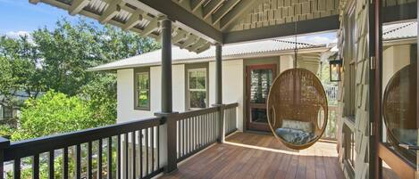 A covered porch with a hanging chair, wooden railing, and a view of a house with a door and windows.