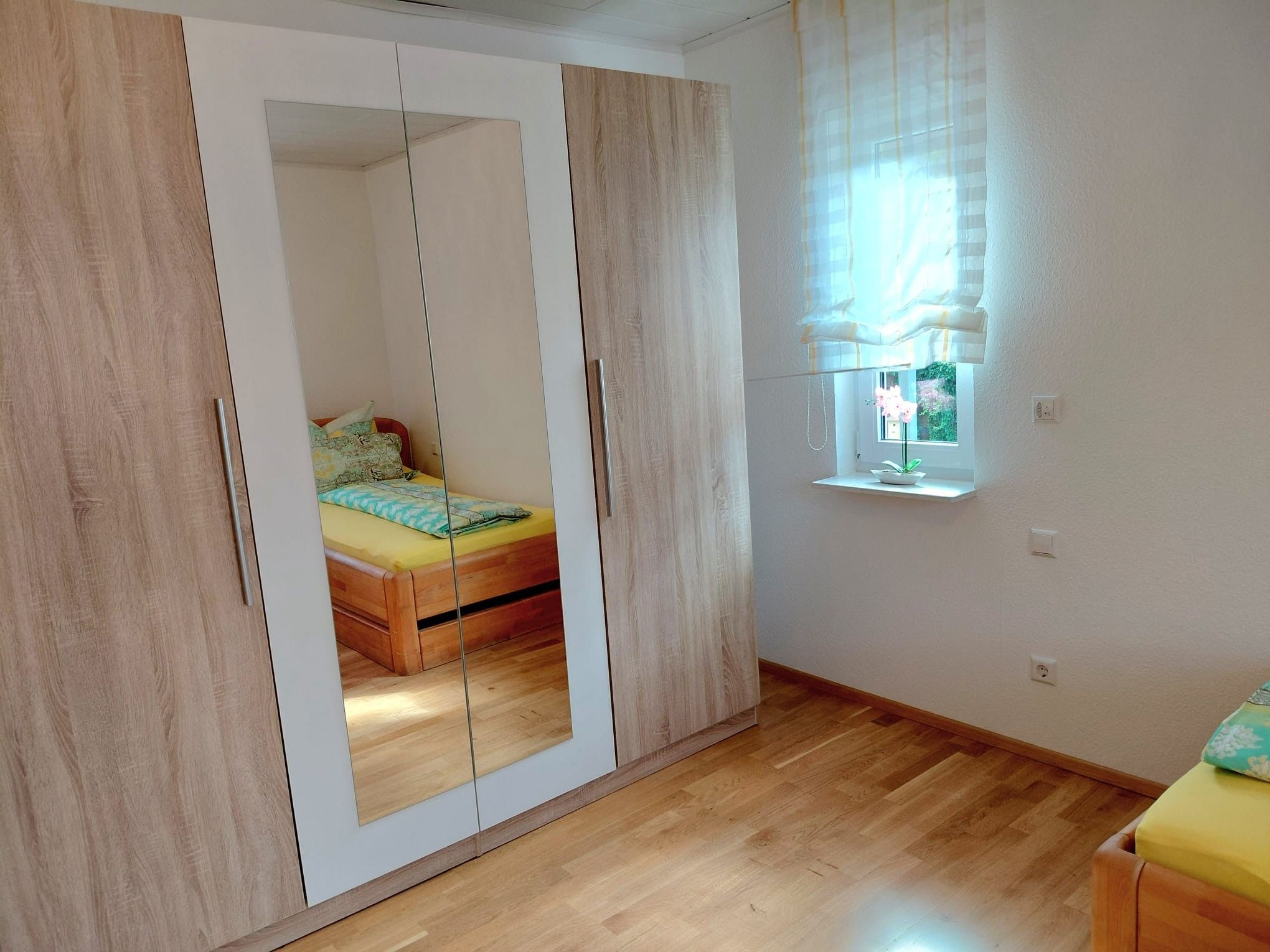 Une chambre avec une armoire en bois, un lit recouvert d’une couverture jaune et une fenêtre donnant sur un paysage verdoyant.