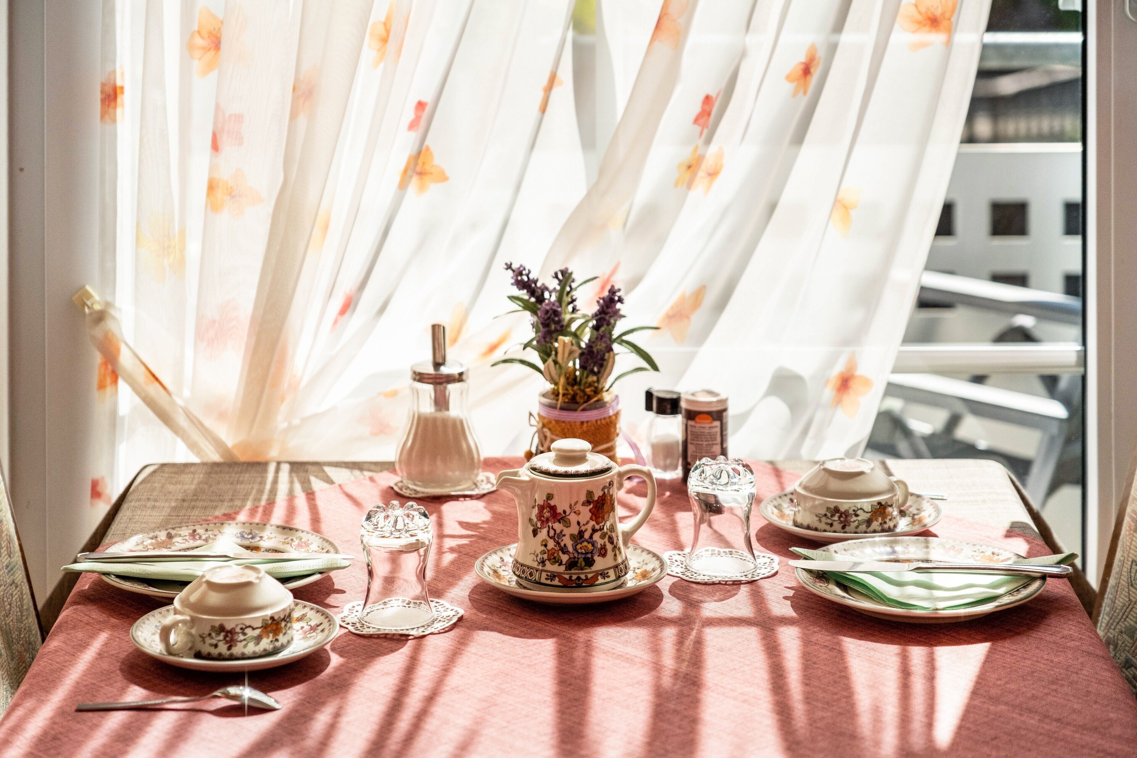 A table set with tea service, a vase with flowers, and a view through sheer curtains.