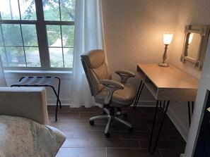 Ein Zimmer mit Schreibtisch, Stuhl und einem Fenster mit Blick ins Grüne.