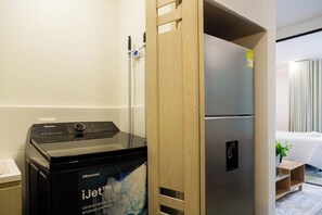 Habitación de hotel moderna y compacta, con lavadora integrada, refrigerador y una cama con edredón blanco.