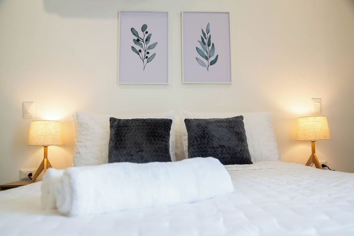 Una cama bien hecha con ropa blanca, dos almohadas oscuras y dos lámparas de noche con pantalla blanca.