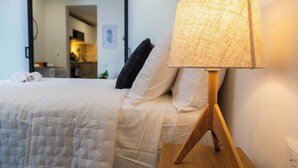 Un lit recouvert d’une couette blanche matelassée, une table de chevet en bois avec une lampe, et une vue sur un coin cuisine.