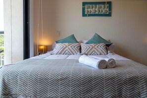 A neatly made bed with patterned bedding and pillows, a wall-mounted artwork, and a window with curtains. - Estudio Boutique con Balcon Vista y Parqueo (Santa Marta)