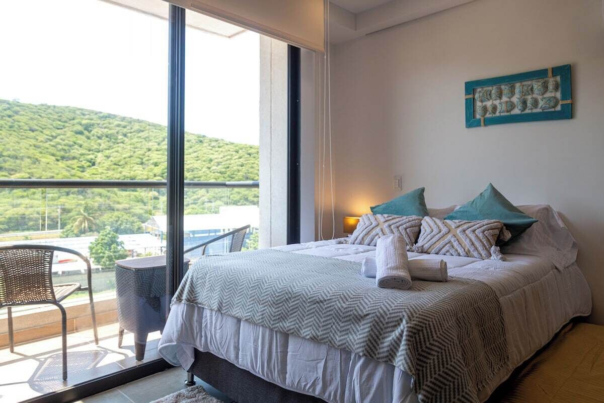 Un dormitorio con un ventanal grande, una cama con muchas almohadas, una mesita pequeña y una silla.