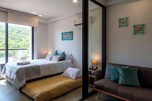 A modern bedroom with a large bed, a bench, a sofa, and a view of greenery through the window. - Estudio Boutique con Balcon Vista y Parqueo (Santa Marta)