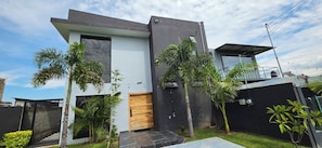 A modern two-story house with a flat roof, a wooden door, and a small garden with palm trees.