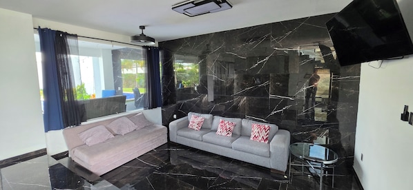 A modern living room with a sofa, a glass coffee table, and a large TV mounted on the wall.