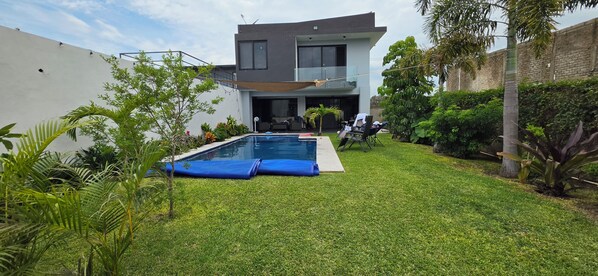 A modern two-story house with a swimming pool, surrounded by well-maintained grass and tropical plants.
