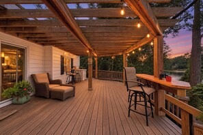 A wooden deck with outdoor furniture, string lights, and a view of a lake at dusk.