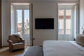 A hotel room with a bed, a chair, a TV, and large windows with a view of buildings. - Príncipe Executive Residence (Madrid)