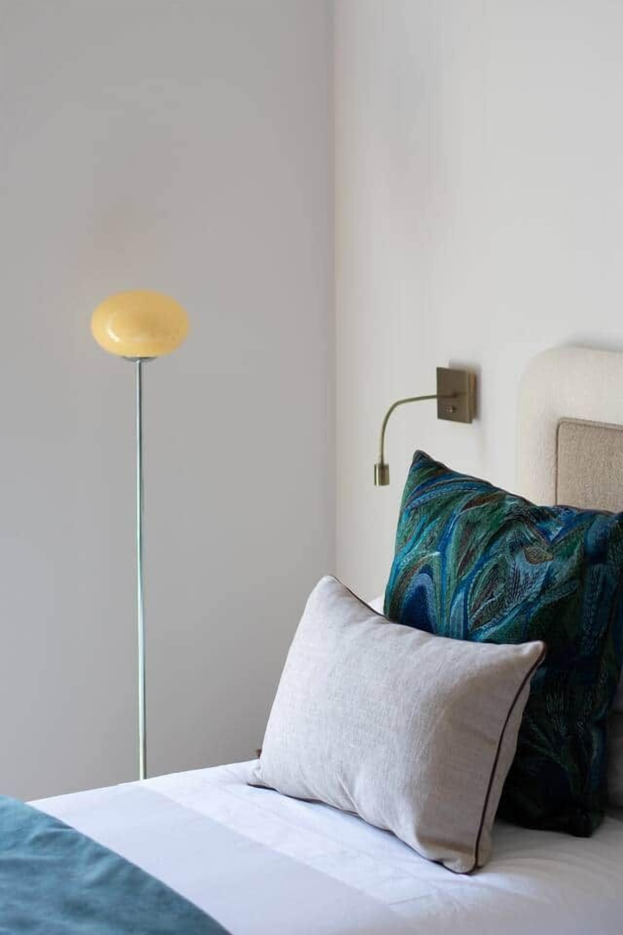 Un letto con una coperta bianca e azzurro mare, un cuscino bianco e un cuscino azzurro con motivo decorativo. Una lampada da terra con paralume giallo è posizionata a sinistra.
