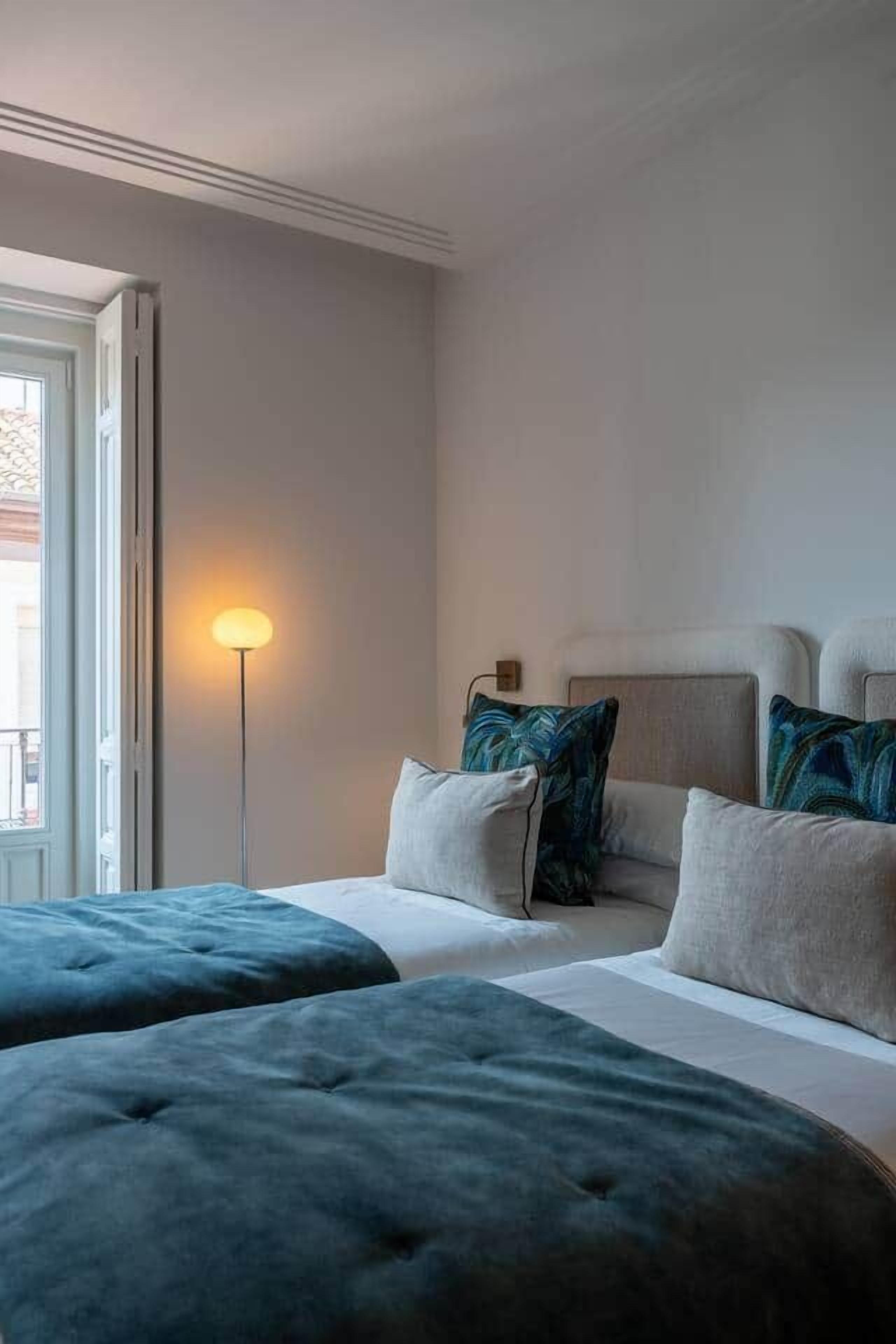 Una habitación de hotel con cama, dos otomanes, una lámpara de pie y una ventana.
