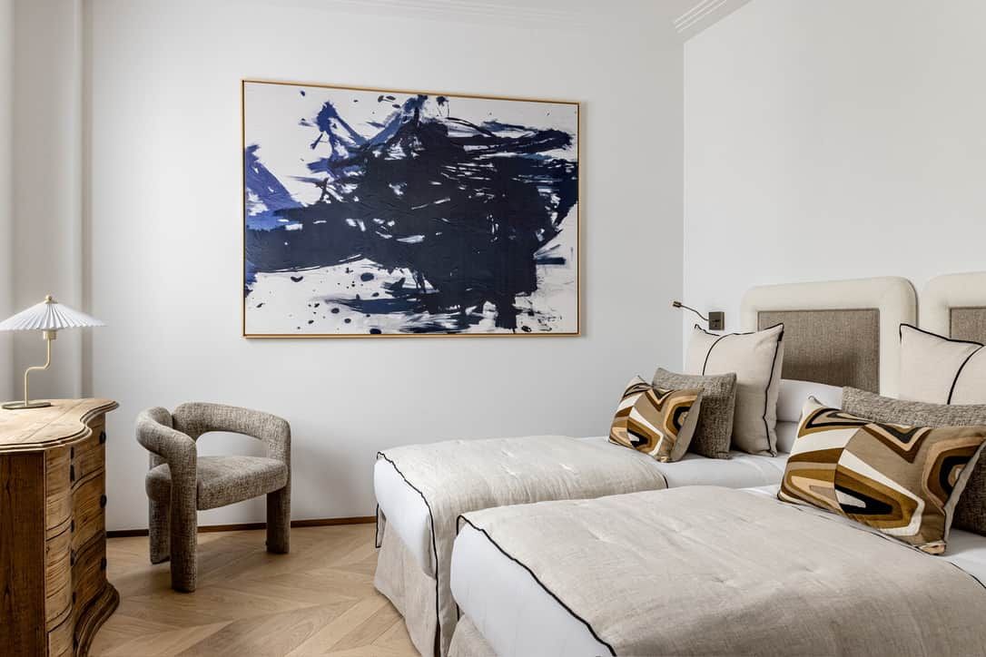 Une chambre avec un grand tableau abstrait, une commode en bois, une chaise et un lit agrémenté d’oreillers à motifs.