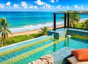 Una piscina con vista a una playa y al océano, rodeada de palmeras y el horizonte de una ciudad.