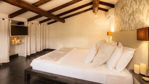 Ein Schlafzimmer mit Holzbalkendecke, einem Bett mit weißer Bettwäsche, einem Nachttisch mit Kerzen und einem an der Wand befestigten Fernseher.