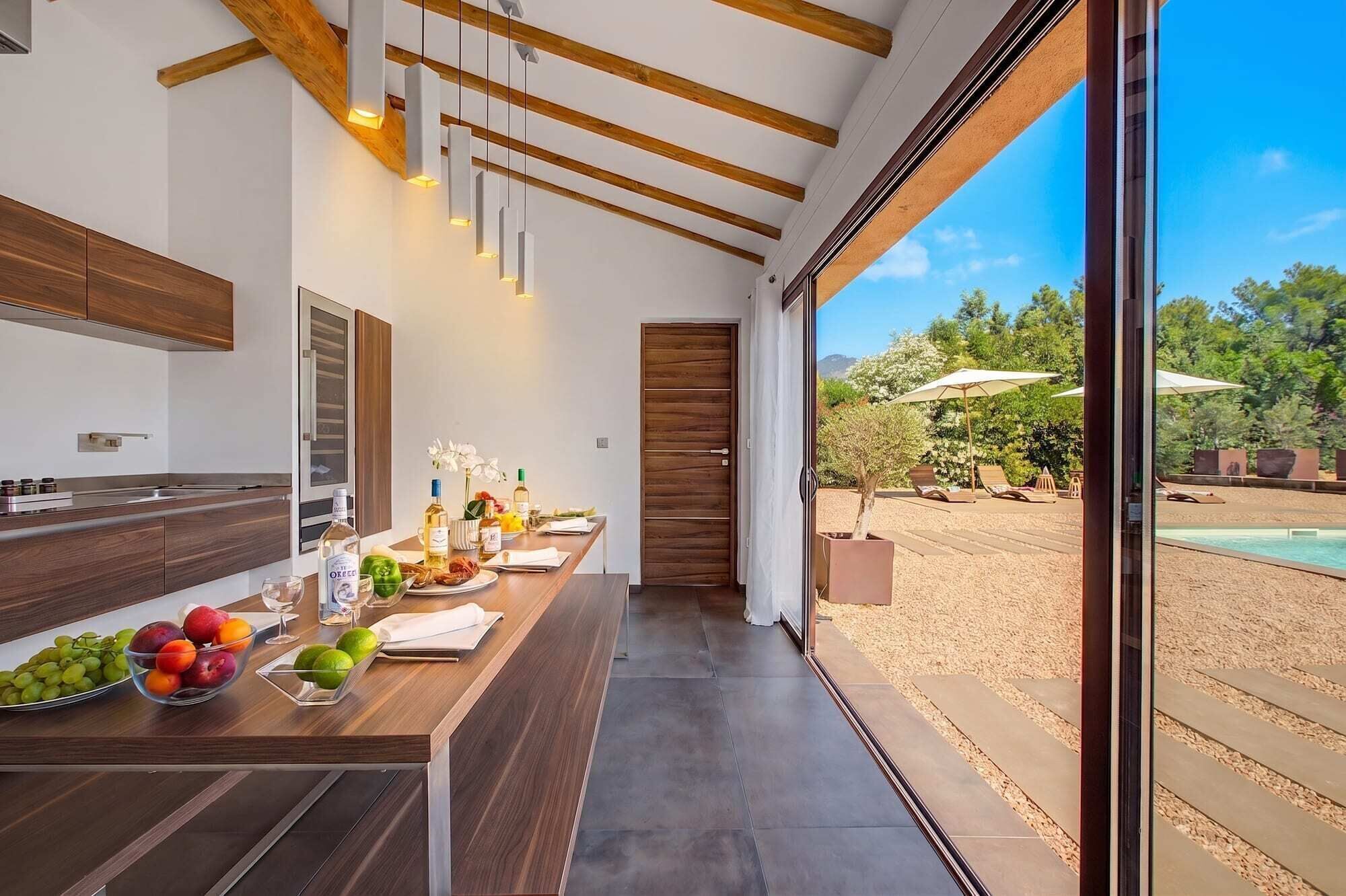 A modern kitchen with a wooden countertop, a dining area with a table set for two, and a view of a pool and garden outside.