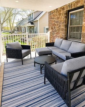 Eine überdachte Terrasse mit einer Steinwand, einem grauen Sofa, zwei Sesseln und einem kleinen runden Tisch.