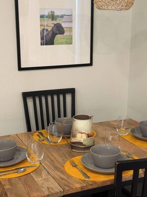 Una mesa de comedor dispuesta con platos grises, cubiertos y vasos, con un cuadro enmarcado encima y una cesta tejida colgada en la pared.